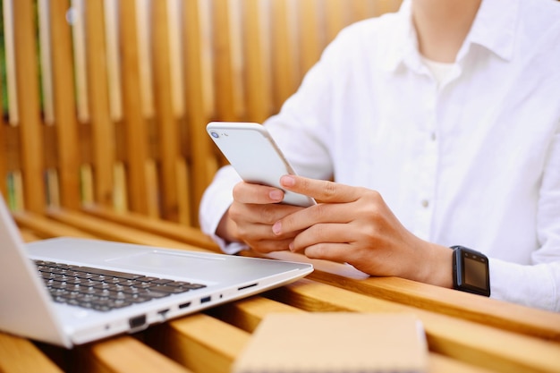 Porträt einer gesichtslosen Frau mit weißem Hemd, die an einem Holztisch in der Nähe eines Laptops sitzt und mit dem Handy im Internet surft, soziale Netzwerke überprüft oder Nachrichten beantwortet