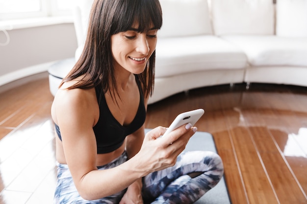 Porträt einer gesunden Frau mit Handy beim Sport oder Yoga auf der Matte im hellen Raum zu Hause