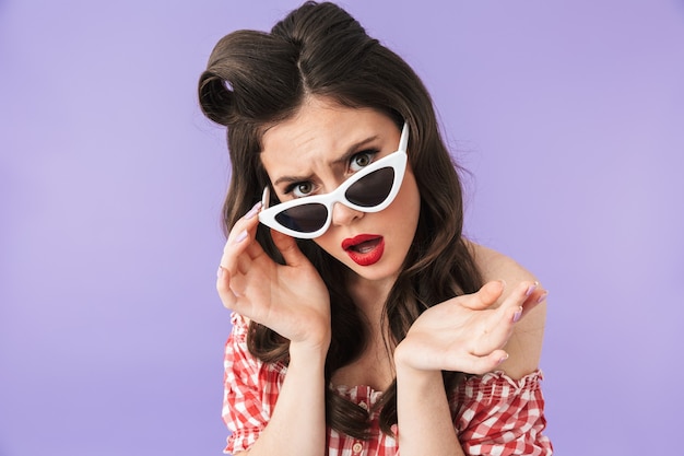 Porträt einer glamourösen Pin-up-Frau im amerikanischen Stil und Retro-Sonnenbrille, die in die Kamera lächelt, isoliert über violetter Wand