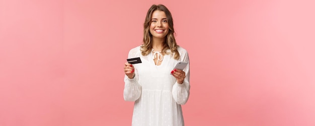 Porträt einer glücklich lächelnden blonden Frau, die Lebensmittel online bestellt, Anwendung zum Einkaufen im Internet-Shop hält, die Kreditkarte und Smartphone hält und erfreut über den rosa Hintergrund der Kamera grinst