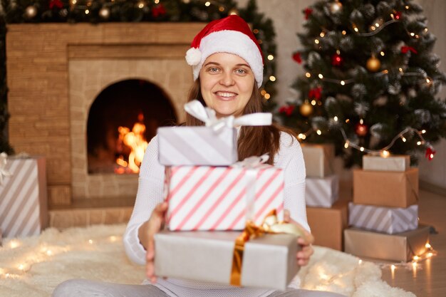 Porträt einer glücklich lächelnden Frau mit rotem Weihnachtsmütze, die einen Stapel von Geschenken mit Blick auf die Kamera hält, Weihnachtsgeschenke feiert und im festlichen Wohnzimmer mit Kamin posiert.