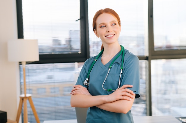 Porträt einer glücklichen ärztin in blaugrüner uniform, die an einem sonnigen tag im büro der medizinischen klinik in der nähe des fensters gekreuzt ist.