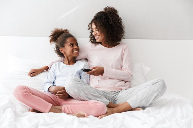 Porträt einer glücklichen afroamerikanischen Frau und ihrer kleinen Tochter, die mit Fernbedienung fernsehen, während sie zu Hause auf dem Sofa sitzt