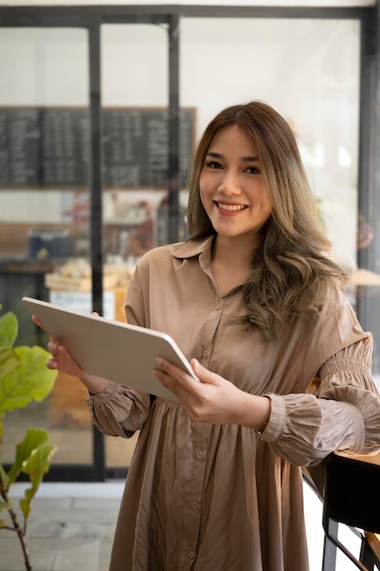 Porträt einer glücklichen asiatischen Besitzerin, die in ihrem Café steht und ein digitales Tablet verwendet