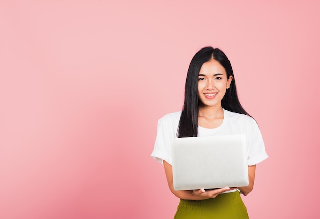 Porträt einer glücklichen asiatischen schönen jungen Frau, die selbstbewusst lächelndes Gesicht hält und einen Laptop verwendet, der zur Kamera schaut, Studioaufnahme isoliert auf rosa Hintergrund, mit Kopierraum