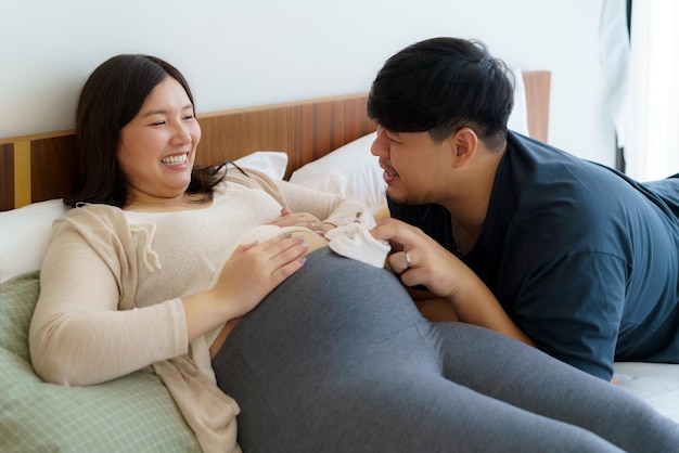 Porträt einer glücklichen asiatischen schwangeren Frau und ihres Mannes, die Babyschuhe auf dem Bett halten