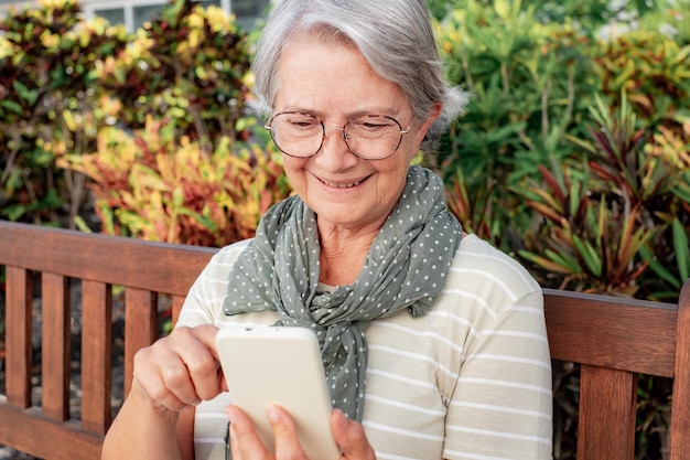 Porträt einer glücklichen, attraktiven Seniorin in T-Shirt und Brille, während sie im öffentlichen Park mit dem Handy im Freien sitzt Lächelnde ältere kaukasische Dame, die Freizeit und Technologie genießt