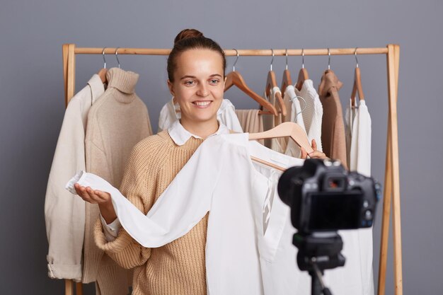 Porträt einer glücklichen, erfreut lächelnden Frau, die in der Nähe eines Kleiderbügels mit Kleidern im Modegeschäft oder zu Hause im Kleiderschrank steht und ihren Anhängern, die Outfits verkaufen, eine weiße Bluse zeigt