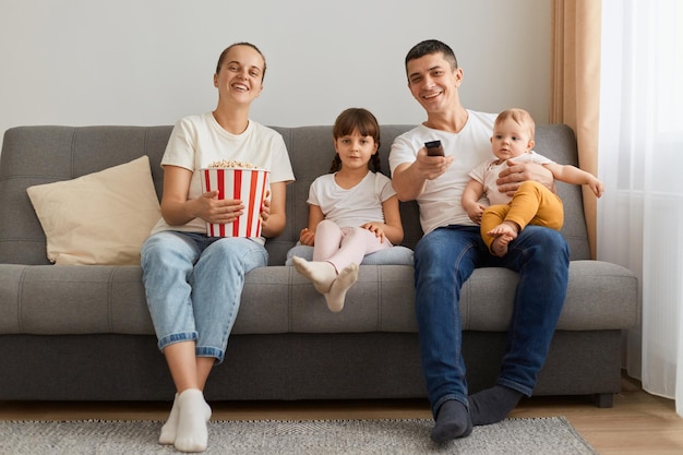 Porträt einer glücklichen Familie, die zu Hause auf einem bequemen Sofa sitzt und fernsieht oder einen Film sieht, der das Glück in Freizeitkleidung ausdrückt und Popcorn und Film genießt