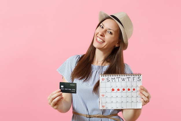 Porträt einer glücklichen Frau im blauen Kleid, Hut mit Kreditkarte, Periodenkalender, Überprüfung der Menstruationstage einzeln auf trendigem rosa Hintergrund. Gynäkologisches Konzept des medizinischen Gesundheitswesens. Platz kopieren.