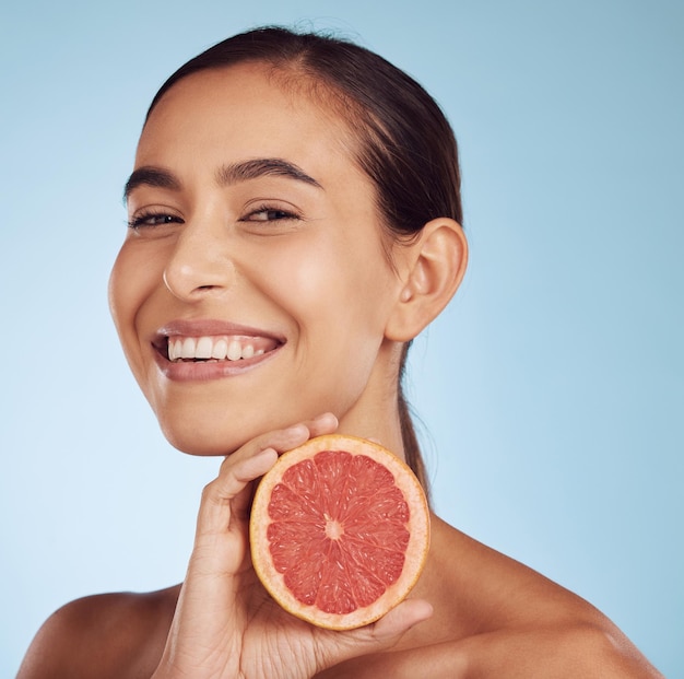 Porträt einer glücklichen Frau mit Hautpflege und Grapefruit für Schönheitskosmetik, Naturprodukt und tropische Gesichtsbehandlung oder Vitamin C. Gesicht einer Person oder eines Modells mit roten Früchten für die Dermatologie auf studioblauem Hintergrund