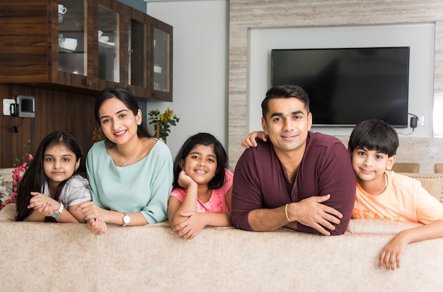 Porträt einer glücklichen indischen asiatischen jungen Familie beim Sitzen auf dem Sofa, auf dem Boden liegend oder an der Wand sitzend