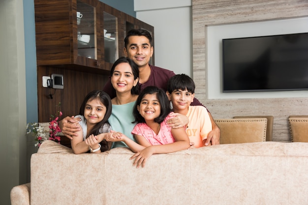 Porträt einer glücklichen indischen asiatischen jungen Familie beim Sitzen auf dem Sofa, auf dem Boden liegend oder an der Wand sitzend