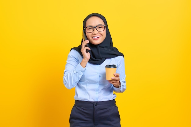 Porträt einer glücklichen jungen asiatischen Frau, die auf dem Handy spricht und eine Tasse Kaffee auf gelbem Hintergrund hält