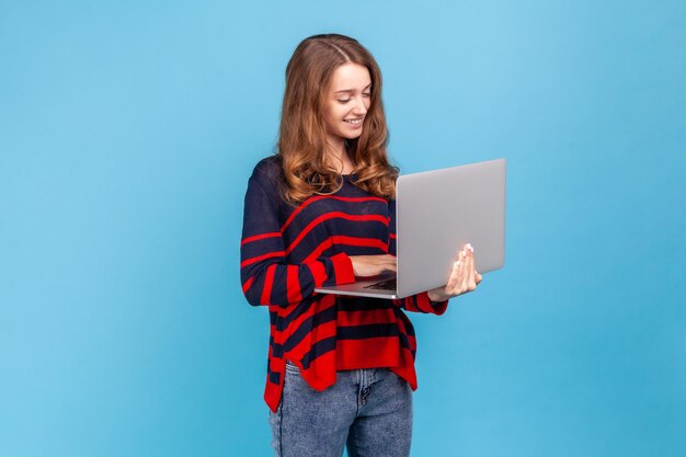 Porträt einer glücklichen jungen erwachsenen Frau, die einen gestreiften Pullover im lässigen Stil trägt, an einem Laptop arbeitet und mit einem charmanten Lächeln auf das Display blickt. Studioaufnahme im Innenbereich isoliert auf blauem Hintergrund.