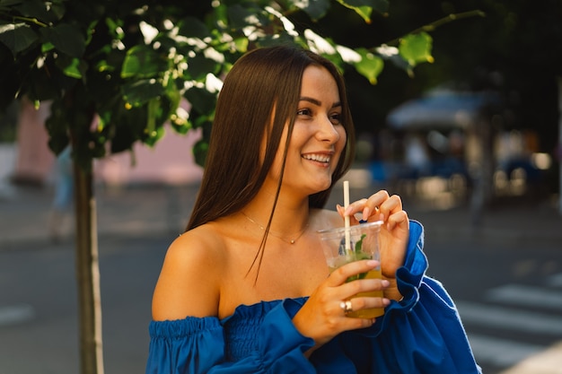 Porträt einer glücklichen jungen Frau, die blaues Kleid trägt und Sommercocktaillimonade trinkt