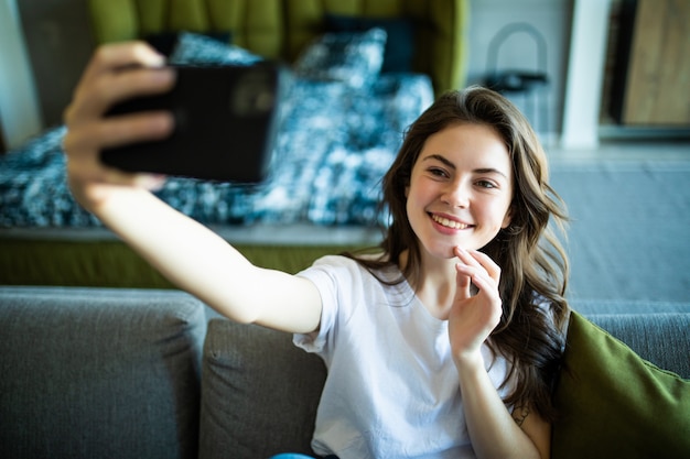 Porträt einer glücklichen jungen Frau, die ein Selfie mit Handy nimmt, während auf einer Couch zu Hause gelehnt