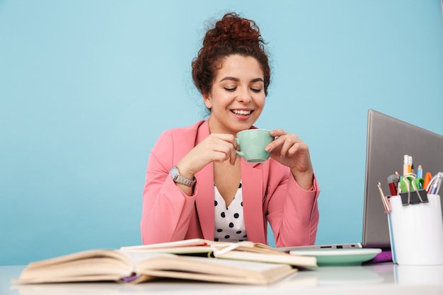 Porträt einer glücklichen jungen Frau, die Kaffee trinkt und lächelt, während sie am Schreibtisch arbeitet, isoliert auf Blau