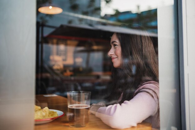 Porträt einer glücklichen jungen Frau durch ein Fenster Sie sitzt am Tisch in einer Bar