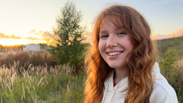 Porträt einer glücklichen jungen Frau mit Brille bei Sonnenuntergang vor natürlichem Hintergrund, lächelt und blickt in die Kamera