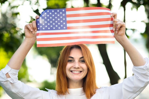 Porträt einer glücklichen jungen rothaarigen Frau, die die USA-Nationalflagge in ihren Händen hält, die im Freien steht