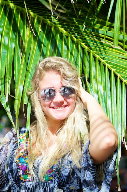 Porträt einer glücklichen jungen schönen Frau mit Sonnenbrille mit tropischer Palme am Strand am Sommertag