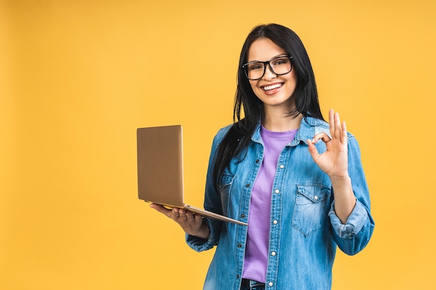 Porträt einer glücklichen jungen, schönen, überraschten Frau mit Brille, die mit einem Laptop steht, der auf gelbem Hintergrund isoliert ist Platz für Text