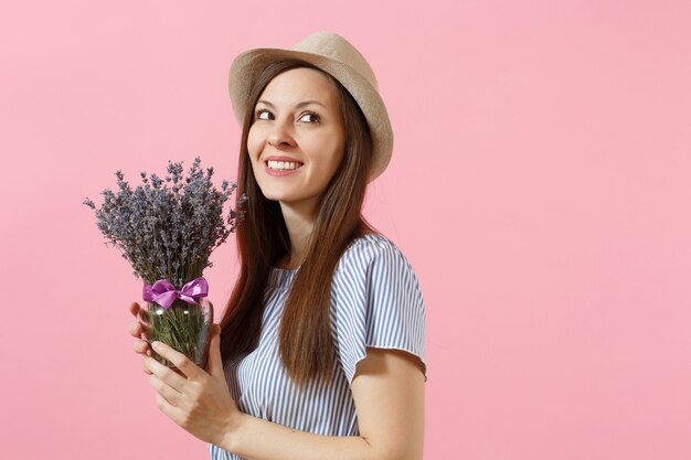 Porträt einer glücklichen jungen, zarten Frau in blauem Strohhut, die einen Strauß schöner lila Lavendelblüten hält, einzeln auf hellem rosafarbenem Hintergrund. Urlaubskonzept zum Internationalen Frauentag