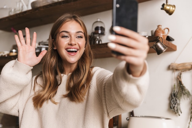 Porträt einer glücklichen kaukasischen Frau in Freizeitkleidung, die Selfie auf dem Smartphone macht und die Hand in der gemütlichen Küche winkt