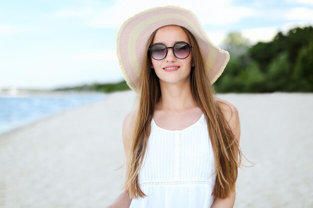 Porträt einer glücklichen lächelnden Frau in freier Glückseligkeit am Strand mit Hut und Sonnenbrille. Ein weibliches Modell in einem weißen Sommerkleid genießt die Natur während der Urlaubsreise