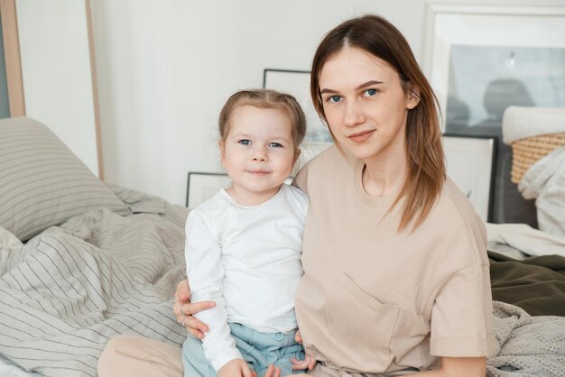 Porträt einer glücklichen, liebevollen alleinerziehenden Mutter, die eine süße kleine Tochter im Vorschulalter umarmt und auf dem Bett sitzt