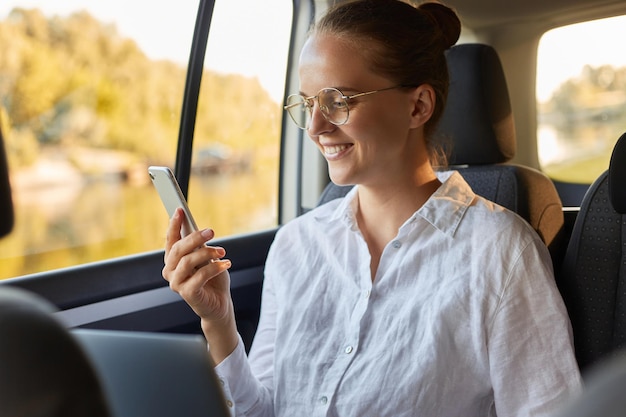 Porträt einer glücklichen optimistischen Frau mit Brille und Brötchenfrisur mit Laptop und Handy, während sie im Auto sitzt und Nachrichten am Telefon liest oder soziale Netzwerke überprüft