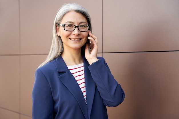 Porträt einer glücklichen reifen Geschäftsfrau mit Brille und klassischer Abnutzung, die am Handy spricht und lächelt, während sie im Freien steht. Geschäftsleute und Kommunikation