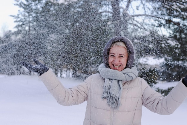 Porträt einer glücklichen, schönen, älteren, pensionierten Frau im Alter, die Spaß mit Schnee im Freien im Wald oder Park am kalten Wintertag hat und lächelnd das Wetter genießt