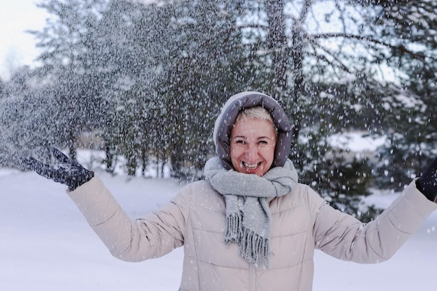 Porträt einer glücklichen, schönen, älteren, pensionierten Frau im Alter, die Spaß mit Schnee im Freien im Wald oder Park am kalten Wintertag hat und lächelnd das Wetter genießt
