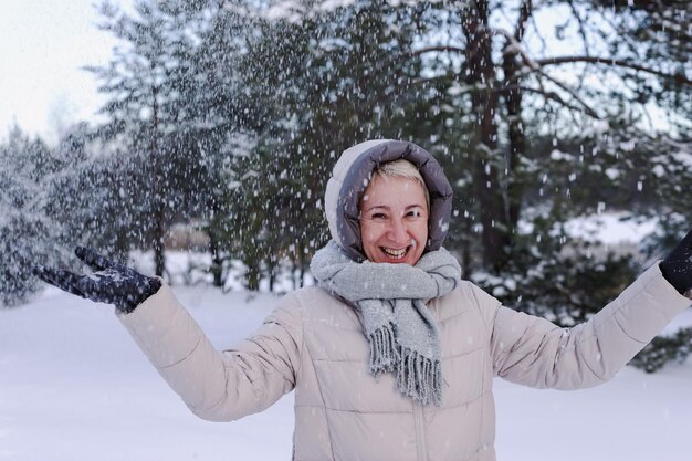 Porträt einer glücklichen, schönen, älteren, pensionierten Frau im Alter, die Spaß mit Schnee im Freien im Wald oder Park am kalten Wintertag hat und lächelnd das Wetter genießt