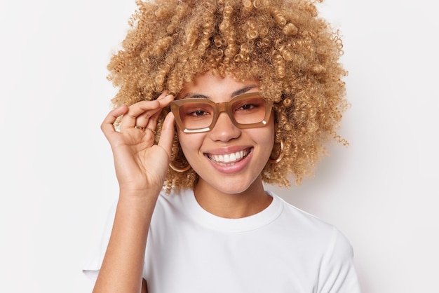 Porträt einer glücklichen, selbstbewussten jungen Frau mit lockigem, buschigem Haar hält die Hand am Brillenrand, lächelt zahnig gekleidet in einem lässigen T-Shirt, das isoliert auf weißem Hintergrund gekleidet ist. Konzept der positiven Emotionen