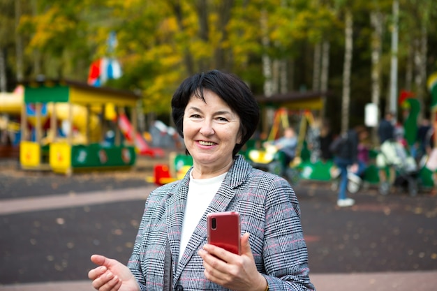 Porträt einer glücklichen Senora für 50 Jahre im Park im Herbst in einer grauen Jacke auf dem Spielplatz. Eine Frau lacht und benutzt ein rotes Smartphone