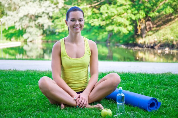 Porträt einer glücklichen sportlichen Frau, die mit einer Yoga-Matte und Wasser im Park sitzt