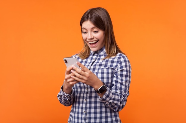 Porträt einer glücklichen, überraschten, brünetten Frau mit charmantem Lächeln, die ein kariertes Hemd trägt, das mit offenem Mund unter Verwendung eines Mobiltelefons steht, Wow-Gesichtsausdruck. Studioaufnahme im Innenbereich isoliert auf orangefarbenem Hintergrund