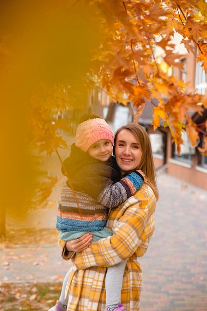 Porträt einer glücklichen und schönen Mutter mit ihrer Tochter in den Armen zwischen Herbstlaub auf der Stadtstr...