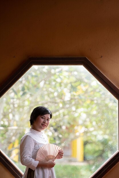 Porträt einer glücklichen vietnamesischen Frau im Ao-Dai-Kleid, die am Fenster steht