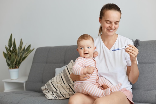 Porträt einer glücklichen, zufriedenen Frau mit weißem T-Shirt im Casual-Stil, die mit ihrer kleinen Tochter und ihrem Schwangerschaftstest in den Händen posiert und positive Emotionen ausdrückt.