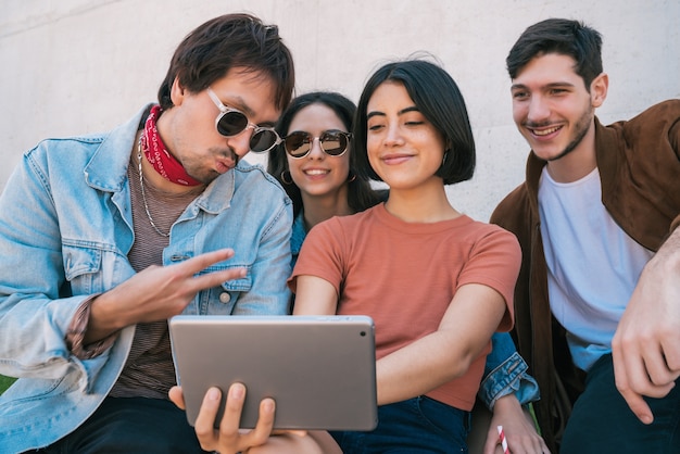 Porträt einer Gruppe von Freunden, die Spaß haben und ein digitales Tablet zusammen benutzen, während sie draußen sitzen. Technologie-, Lifestyle- und Freundschaftskonzept.