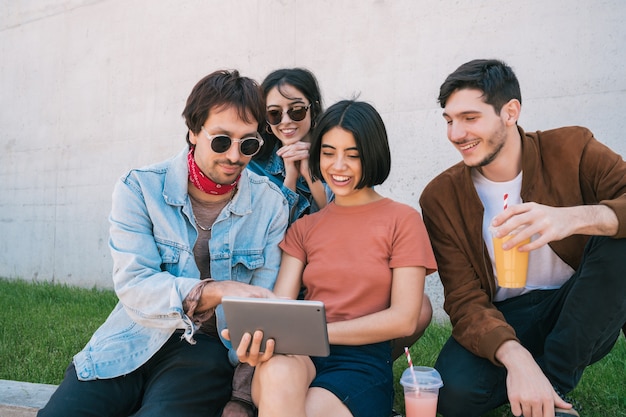 Porträt einer Gruppe von Freunden, die Spaß haben und ein digitales Tablet zusammen benutzen, während sie draußen sitzen. Technologie-, Lifestyle- und Freundschaftskonzept.