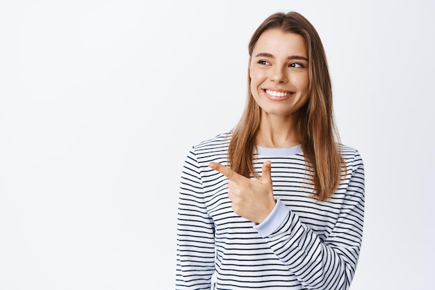 Porträt einer gut aussehenden jungen Frau mit weißem Lächeln, die nach links zeigt und zufrieden aussieht, ihre Wahl im Geschäft trifft und den Produktartikel zeigt, weiß