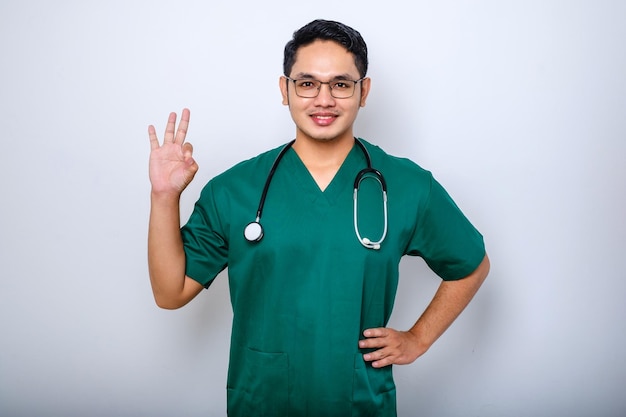 Porträt einer gutaussehenden asiatischen Arztkrankenschwester in Scrubs und Stethoskop, die positiv lächelt und mit der Hand und dem Finger einen erfolgreichen Ausdruck macht