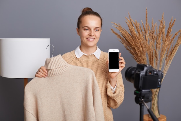 Porträt einer gutaussehenden Frau mit Brötchenfrisur, die ein Video vor der Kamera aufnimmt, mit einem Stativ, das einen warmen, weichen Pullover und ein Smartphone mit leerem Display für den Werbebereich zeigt