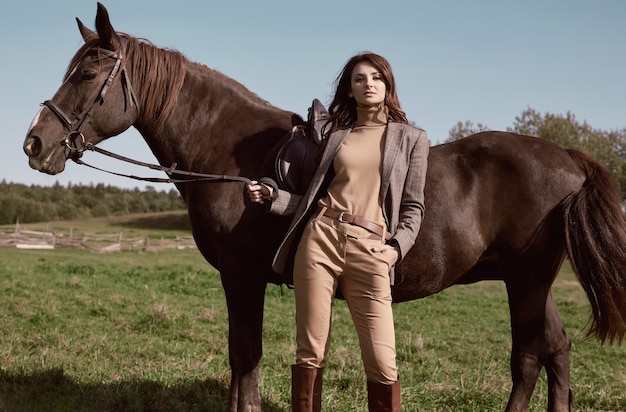 Porträt einer herrlichen brünetten Frau in einer eleganten karierten braunen Jacke, die mit einem Pferd auf Landlandschaft aufwirft