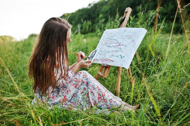 Porträt einer herrlichen glücklichen jungen Frau im schönen Kleid, das auf dem Gras sitzt und auf Papier mit Aquarellen malt.